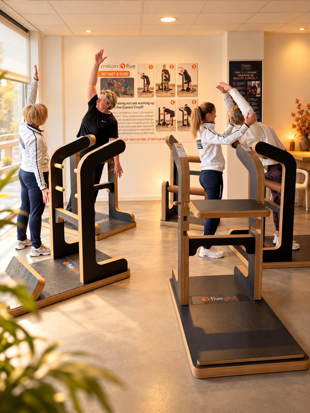 Vier leden doen rekoefeningen op de MILON FIVE-toestellen in een lichte trainingsruimte, begeleid door een coach in een witte jas