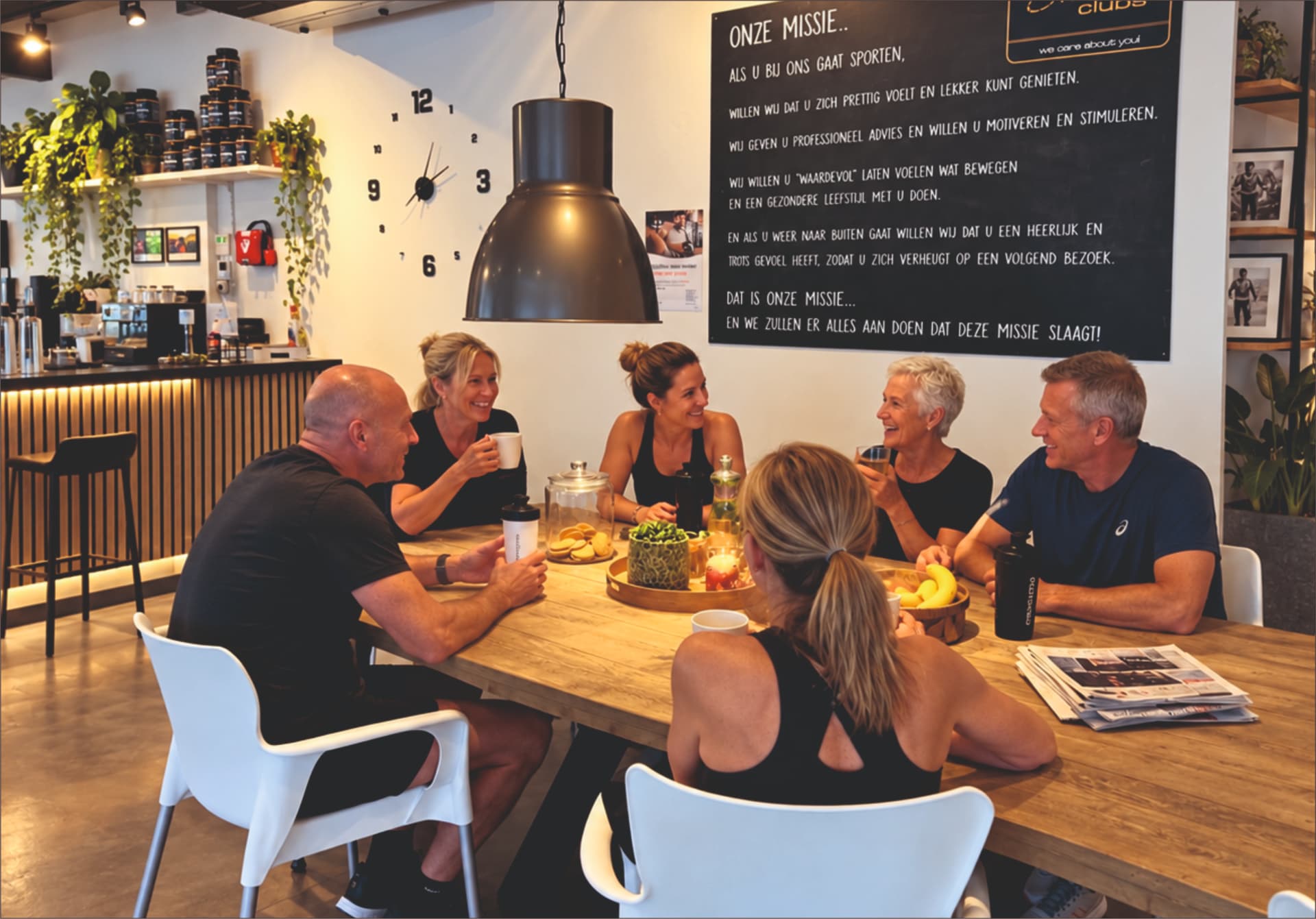Zes leden en coaches zitten lachend met drankjes aan de tafel in de club, met het bord met de missie van Corpus Milon Clubs aan de wand