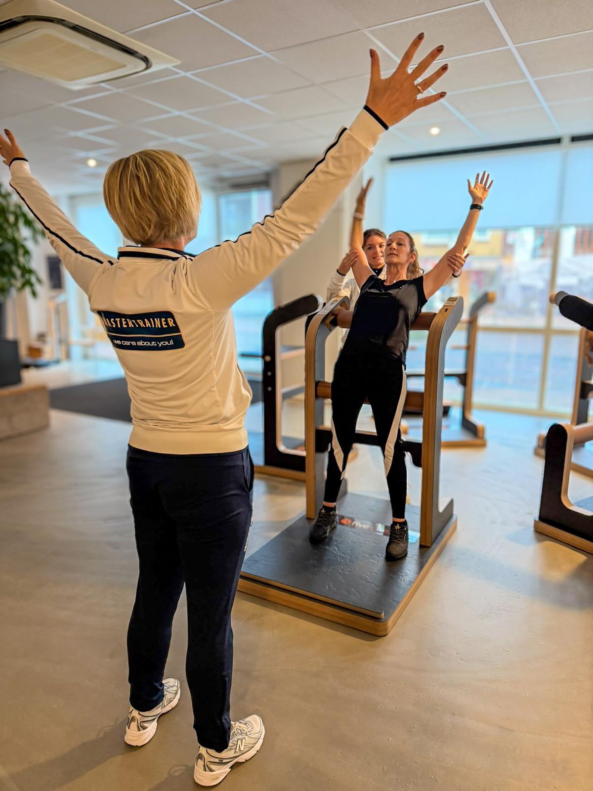Een lid staat met beide armen omhoog op een MILON FIVE-toestel terwijl een coach in een witte jas haar vanaf de voorgrond begeleidt en een tweede coach haar vanuit de rug ondersteunt