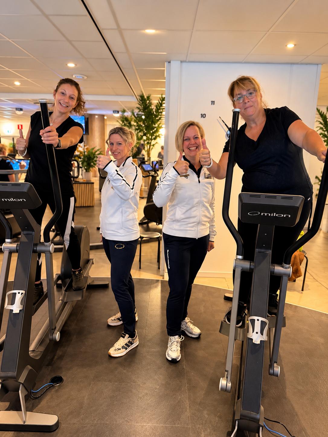 Vier glimlachende leden steken hun duim op; twee van hen staan op een Milon-crosstrainer en twee staan ertussen, in een lichte clubruimte